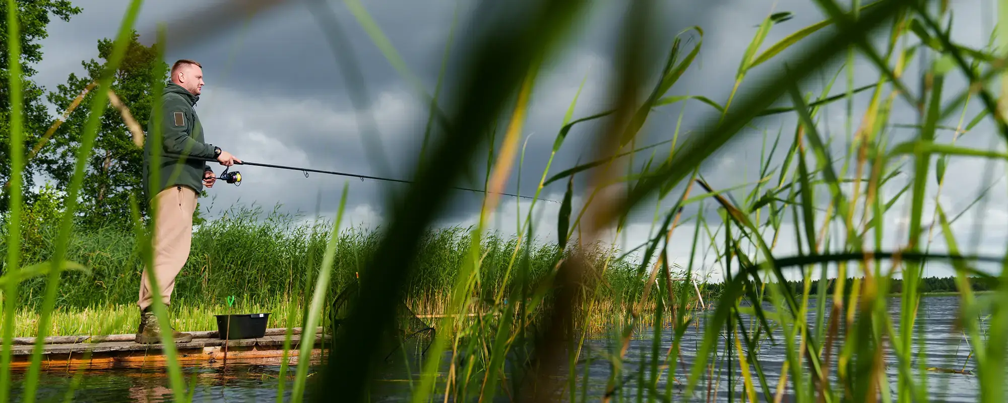 Angler steht mit ausgeworfener Angel auf einem Steg umgeben von Schilf